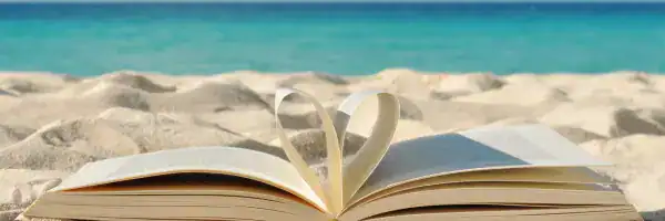 Ein aufgeschlagenes Buch liegt im Sand am Strand, die Seiten sind zu einem Herz geformt, im Hintergrund das Meer.