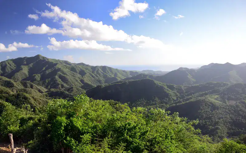 Weite Aussicht über die grünen Bergketten der Sierra Maestra in Kuba mit Blick bis zum Meer unter blauem Himmel mit einzelnen Wolken.