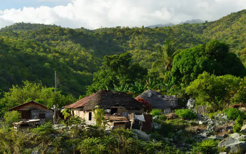 Kleine Holzhäuser inmitten dichter grüner Vegetation am Hang der Sierra Maestra, im Hintergrund bewaldete Berge unter leicht bewölktem Himmel.