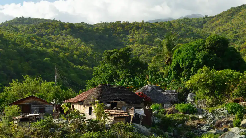 Kleine Holzhäuser inmitten dichter grüner Vegetation am Hang der Sierra Maestra, im Hintergrund bewaldete Berge unter leicht bewölktem Himmel.