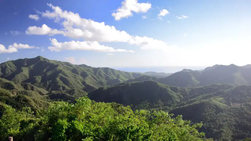 Weite Aussicht über die grünen Bergketten der Sierra Maestra in Kuba mit Blick bis zum Meer unter blauem Himmel mit einzelnen Wolken.