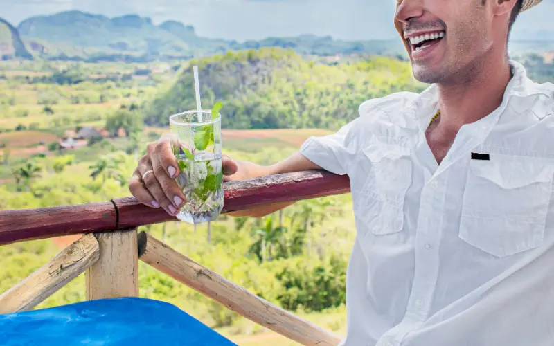 Lachender Mann mit Mojito in der Hand, im Hintergrund die grüne Landschaft des Viñales-Tals in Kuba