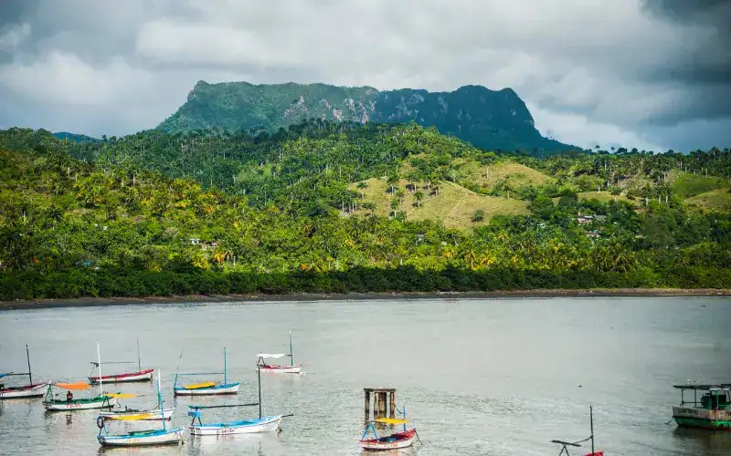 Mehrere bunte Fischerboote liegen im Wasser vor der Küste, dahinter erheben sich grüne Hügel mit dem markanten Tafelberg El Yunque (Kuba) unter bewölktem Himmel.