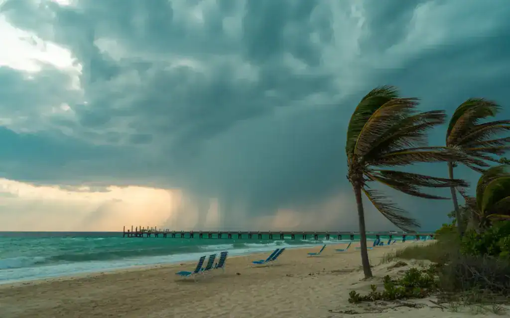 Ein leerer Strand in Varadero mit windgebeugten Palmen und grauen Wolken am Himmel, im Hintergrund zieht ein tropischer Regenschauer über das Meer.